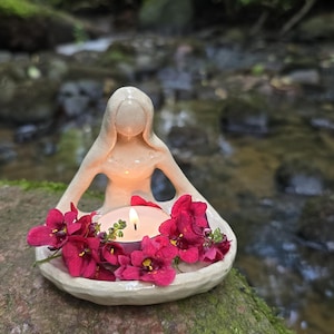 Op de afbeelding: Een keramische kandelaar in de vorm van een zittende figuur, met een brandende kaars omringd door levendige rode bloemen. De houder staat op een met mos bedekte rots, met een vage beek op de achtergrond.
