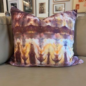 May include: A brown, yellow and purple tie-dye pillow with a white background. The pillow is sitting on a light brown sofa.