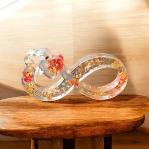 Könnte beinhalten: Ein klares Unendlichkeitszeichen aus Harz mit Blattgold und roten floralen Akzenten. Die Skulptur steht auf einem Holztisch und zeigt ihr kompliziertes Design und ihre künstlerischen Details. Das Symbol steht für Ewigkeit und Liebe.