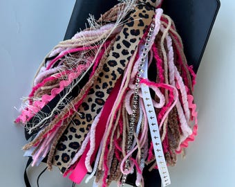 May include: A black journal with a decorative tassel. The tassel features a leopard print ribbon, pink and tan yarn, and a beaded strand. A small pink pom-pom and a white ribbon with crosses are also included.