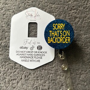 Könnte beinhalten: Ein Ausweis-Rollhalter mit einem blauen, glitzernden, kreisförmigen Design mit dem gelben Text "SORRY THAT'S ON BACKORDER". Der Ausweis-Rollhalter ist an einer weißen Karte mit dem Text "Shady Lake Designs" und "Find us on ebay" befestigt.