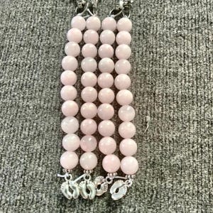 Puede incluir: Tres pulseras de cuentas rosas claras con cierres y dijes plateados. Los dijes tienen forma de pequeños pies. Las cuentas son redondas y parecen estar hechas de un material liso y pulido.