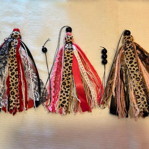 Puede incluir: Tres borlas decorativas con una mezcla de telas, incluyendo estampado de leopardo, encaje y cintas. Cada borla tiene una parte superior con cuentas y una cuerda para colgar. Los colores incluyen negro, rojo, blanco, rosa y marrón.