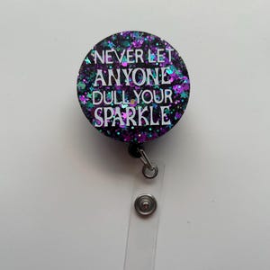 Op de afbeelding: Een zwarte en paarse glitter badgehouder met een witte tekst overlay die "Laat nooit iemand je glans dof maken" luidt.