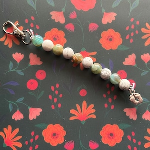 Puede incluir: Un llavero de plata con un dije de dos pequeñas huellas de pies. La cadena está hecha de cuentas verdes, blancas y marrones.