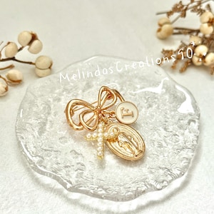 May include: Gold-toned brooch featuring a bow design, a circular charm with the letter "F", a cross adorned with pearls, and a religious medallion. The brooch is displayed on a clear, textured surface. The text "Melindas Creations 50" is visible.