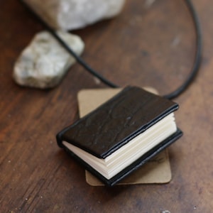 Può includere: Un ciondolo a forma di libro in miniatura rilegato in pelle nera con pagine bianche, sospeso a un cordino nero. Il piccolo libro è appoggiato su un'etichetta marrone, con due pietre bianche sullo sfondo. Il ciondolo è su una superficie di legno.
