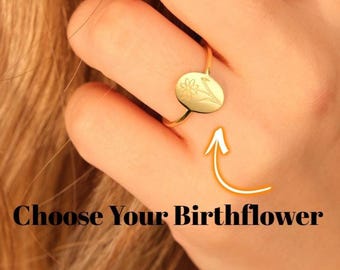 May include: A gold-colored ring with an oval-shaped face featuring a floral design. The ring is on a finger, with the text "Choose Your Birthflower" below it, accompanied by an arrow pointing to the ring.