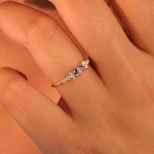May include: A gold ring featuring a central oval-cut purple gemstone, flanked by smaller, clear gemstones. The ring is set on a delicate gold band, creating an elegant and refined aesthetic. The ring is a piece of jewelry.