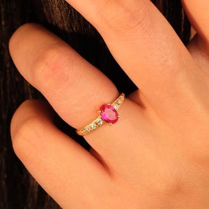 May include: A gold ring featuring a large oval ruby gemstone as the centerpiece. The band is also gold and has small, clear accent stones set along the sides. The ring is on a finger, with the hand in the frame.