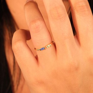 May include: A gold-colored ring with an open design, featuring a blue and a light green rectangular gemstone. The ring is worn on a finger, showcasing the jewelry's delicate details and color contrast.
