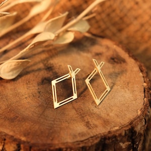 May include: A pair of gold-colored geometric earrings. Each earring features a diamond-shaped design with a smaller, open diamond inside. The earrings are set against a rustic, wooden background with dried foliage.