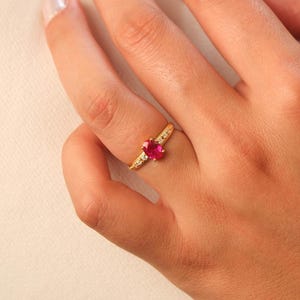 May include: A gold ring featuring a heart-shaped, bright red gemstone as the centerpiece. The band is also gold, with small, clear accent stones set along the sides. The ring is shown on a person's finger.