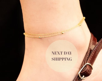 May include: A delicate gold-colored anklet with two strands and small beaded accents. The anklet is worn on an ankle, paired with a brown sandal with a silver buckle. The image has a black background.
