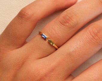 May include: A gold ring with an open design, featuring a blue and a green rectangular gemstone. The ring is worn on a finger, showcasing the delicate craftsmanship and vibrant colours of the gemstones.