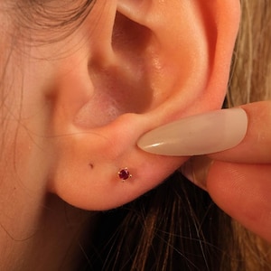 Może przedstawiać: Zbliżenie złotego kolczyka sztyftu z małym, okrągłym, czerwonym kamieniem szlachetnym. Kolczyk znajduje się w uchu osoby. Palec dotyka ucha.