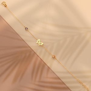 May include: A delicate gold bracelet featuring a butterfly charm and small heart and purple gemstone accents. The bracelet is displayed on a neutral background, showcasing its fine details and elegant design. The bracelet is a beautiful piece of jewelry.