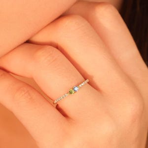 May include: A delicate gold ring featuring a row of small, clear stones. The ring has a cluster of three gemstones: a light green, a light blue, and a clear stone. The ring is on a person's finger.