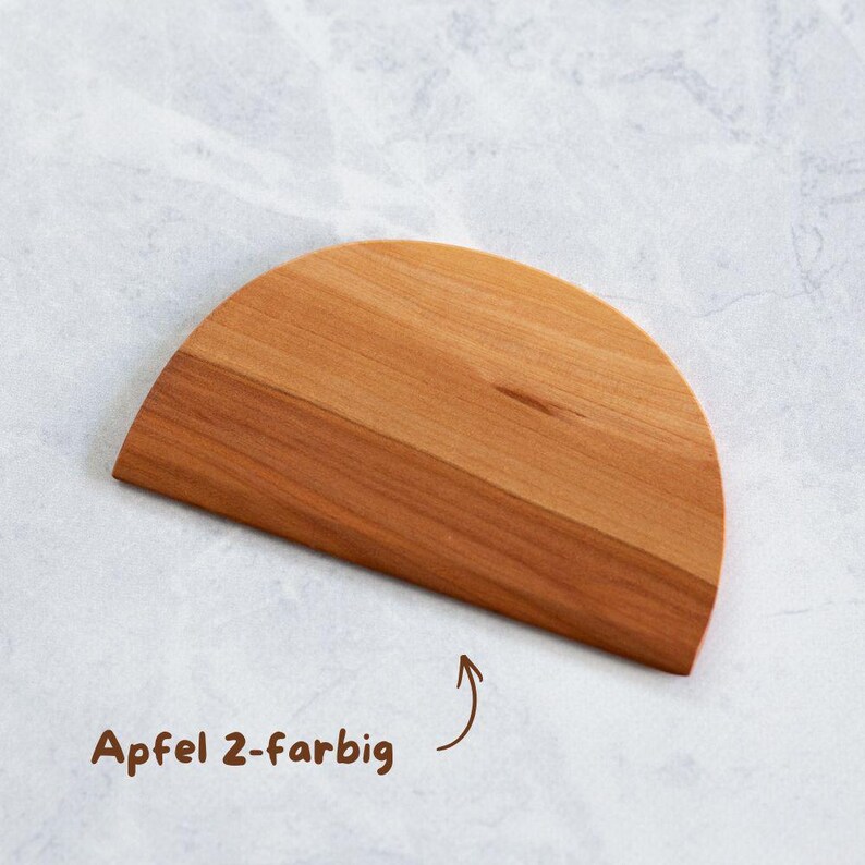 Ein halbkreisförmiger Teigschaber aus Holz in hellbrauner Farbe. Der Text "Apfel 2-farbig" befindet sich in der unteren linken Ecke. Der Teigschaber ist aus Holz und hat eine glatte Oberfläche.
