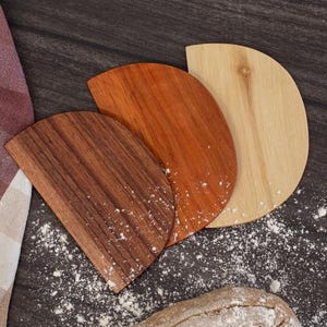 Könnte beinhalten: Drei Teigschaber aus Holz in verschiedenen Braun- und Beigetönen sind auf einer dunklen, mit Mehl bestäubten Oberfläche angeordnet. Links ist ein kariertes Tuch zu sehen, unten ein Brot.
