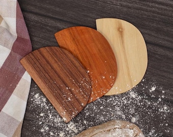 May include: Three wooden dough scrapers in varying shades of brown and tan are arranged on a dark surface dusted with flour. A checkered cloth is visible on the left, and a loaf of bread is at the bottom.