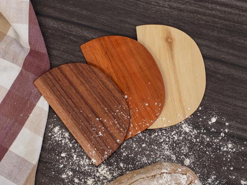 Drei Teigschaber aus Holz in verschiedenen Braun- und Beigetönen sind auf einer dunklen, mit Mehl bestäubten Oberfläche angeordnet. Links ist ein kariertes Tuch zu sehen, unten ein Brot.