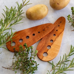 Könnte beinhalten: Zwei Kochlöffel aus Holz mit mehreren Löchern, zusammen mit frischem Rosmarin, Thymian und zwei Kartoffeln. Die Löffel sind hellbraun und haben eine glatte Oberfläche.