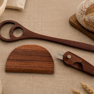 Könnte beinhalten: Ein dunkelbraunes Brotmesser aus Holz mit Metallklinge, ein halbkreisförmiger Teigschaber aus Holz und ein langer Brotwirbler aus Holz mit zwei kreisförmigen Öffnungen am Griffende, alle aus glattem, poliertem Holz mit sichtbarer Maserung.