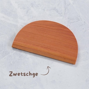 K&ouml;nnte beinhalten: Ein halbkreisf&ouml;rmiges Schneidebrett aus Holz mit rotbraunem Finish. Das Brett hat eine flache Unterkante und eine gebogene Oberseite. Das Wort "Zwetschge" ist in brauner Schrift neben dem Brett geschrieben.