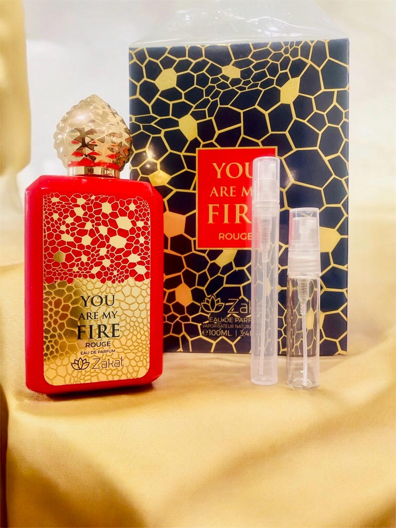 Puede incluir: Un frasco de perfume rojo de vidrio con una tapa y una etiqueta doradas. El frasco est&aacute; etiquetado con "You Are My Fire Rouge Eau de Parfum Zakat". El frasco est&aacute; sobre una superficie dorada con dos peque&ntilde;os frascos pulverizadores transparentes.