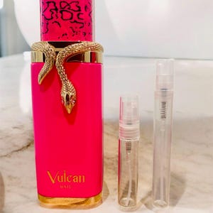 May include: A bright pink perfume bottle with a gold snake wrapped around it. The bottle has a pink and black patterned cap and the words "Vulcan BAIE" in gold. Two small, clear spray bottles are next to it.