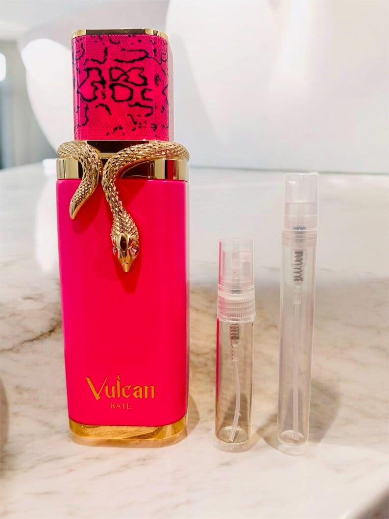 May include: A bright pink perfume bottle with a gold snake wrapped around it. The bottle has a pink and black patterned cap and the words "Vulcan BAIE" in gold. Two small, clear spray bottles are next to it.