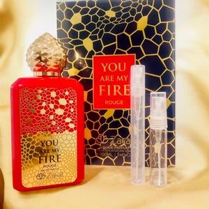 Puede incluir: Una botella de perfume de vidrio rojo con una tapa dorada y una etiqueta dorada que dice "You Are My Fire Rouge" y un logotipo de flor de loto dorada. La botella est&aacute; sobre una superficie dorada con dos peque&ntilde;as botellas de vidrio transparente con pulverizadores.