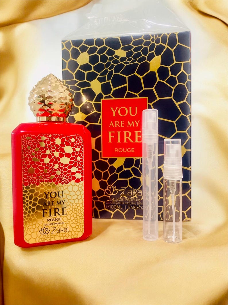 Puede incluir: Una botella de perfume de vidrio rojo con una tapa dorada y una etiqueta dorada que dice "You Are My Fire Rouge" y un logotipo de flor de loto dorada. La botella est&aacute; sobre una superficie dorada con dos peque&ntilde;as botellas de vidrio transparente con pulverizadores.