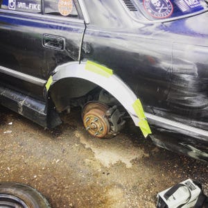 May include: A close-up of a black car's rear wheel well with a new silver fender attached with yellow tape. The rusty brake rotor and caliper are visible. The car has stickers on the window.