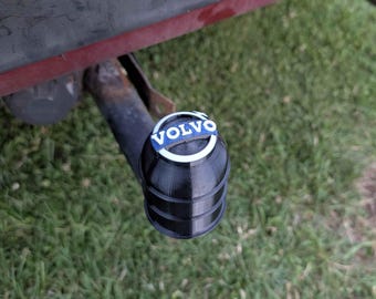 May include: A black trailer hitch cover with the Volvo logo in white and blue. The cover is cylindrical with horizontal ridges. The background is green grass.
