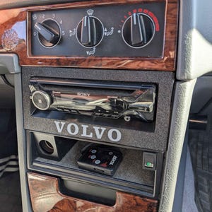May include: A close-up of a car's dashboard with a woodgrain trim. The dashboard features a Sony radio, a Volvo logo, and a black box with a green light. The dashboard also has a set of controls for the car's climate system.