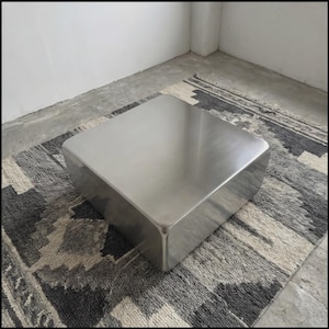 Può includere: Un tavolino quadrato moderno in acciaio inossidabile con angoli arrotondati. Il tavolo è posto su un tappeto grigio e bianco a fantasia. La stanza ha un'estetica minimalista con pavimento in cemento e pareti bianche.