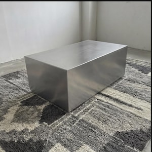 Może przedstawiać: Duży, prostokątny stolik kawowy ze szczotkowanego metalu. Stół jest solidnym blokiem metalu z matowym wykończeniem. Stoi na wzorzystym szaro-białym dywanie. Stół ma około 122 cm długości, 61 cm szerokości i 41 cm wysokości.