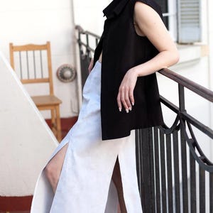 May include: A sleeveless black top and a white skirt with a high slit are worn with black platform sneakers. The outfit is styled on a balcony with a metal railing and a wooden chair in the background.