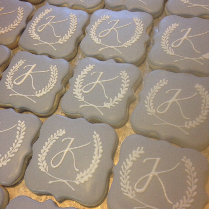 May include: A collection of square-shaped sugar cookies decorated with a white icing design of a letter "J" inside a wreath. The cookies are a light blue color.