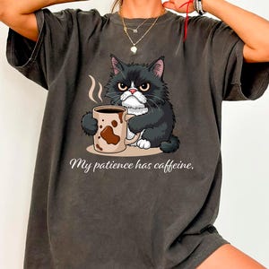 Puede incluir: Camiseta gris oscuro con un gato gruñón blanco y negro sosteniendo una taza de café. La taza tiene un diseño marrón. El texto "My patience has caffeine" está debajo de la ilustración del gato.