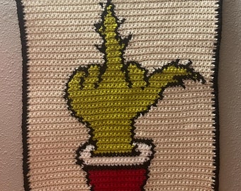 May include: A crocheted wall hanging featuring a green middle finger in a red pot. The hanging is framed with black yarn and has a white background.