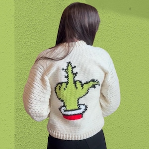 May include: A cream-coloured long-sleeve crochet jumper featuring a large graphic on the back. The design depicts a bright green hand with a black outline making a middle finger gesture, emerging from a white cuff with a red band at the bottom.