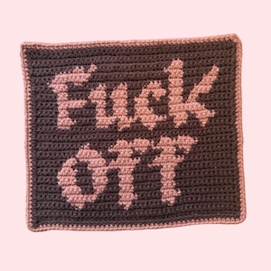 Puede incluir: Un rectángulo tejido a ganchillo con las palabras "Fuck Off" en rosa claro sobre un fondo gris oscuro. El borde es de un rosa claro a juego. El artículo hecho a mano se muestra sobre un fondo rosa claro.