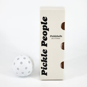 Può includere: Una pallina da pickleball bianca con fori grigi è accanto a una scatola color crema. La scatola ha le parole "Pickle People" in grandi lettere nere e "Pickleballs Set of three" in testo nero più piccolo.