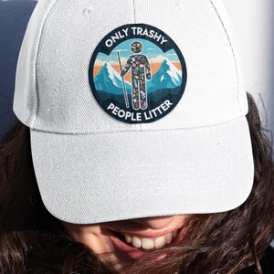May include: White baseball cap with a circular patch featuring a person with a staff in front of mountains and the text "ONLY TRASHY PEOPLE LITTER". The cap is made of a durable fabric.
