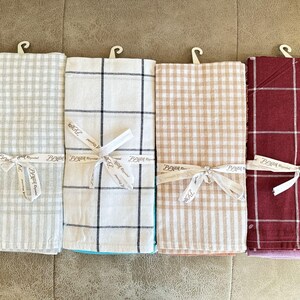 Könnte beinhalten: Vier Küchentücher mit verschiedenen Mustern. Die Tücher sind weiß mit grauem, schwarzem, beigefarbenem und rotem Gingham-Muster. Jedes Tuch hat ein weißes Band, das um es herum gebunden ist, mit dem Text "Beyond Blessed", der auf das Band gedruckt ist.