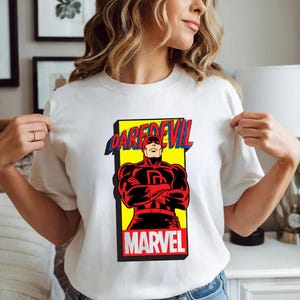 May include: White t-shirt featuring a graphic of the Marvel Comics character Daredevil. The design includes the character in a red and black suit, the word "DAREDEVIL" in blue and red, and the word "MARVEL" in white.