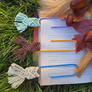 May include: Three handmade macrame bookmarks in shades of blue, brown, and cream, each with a different color of plastic clip. The bookmarks are resting on the pages of a book, with grass and fall foliage in the background.